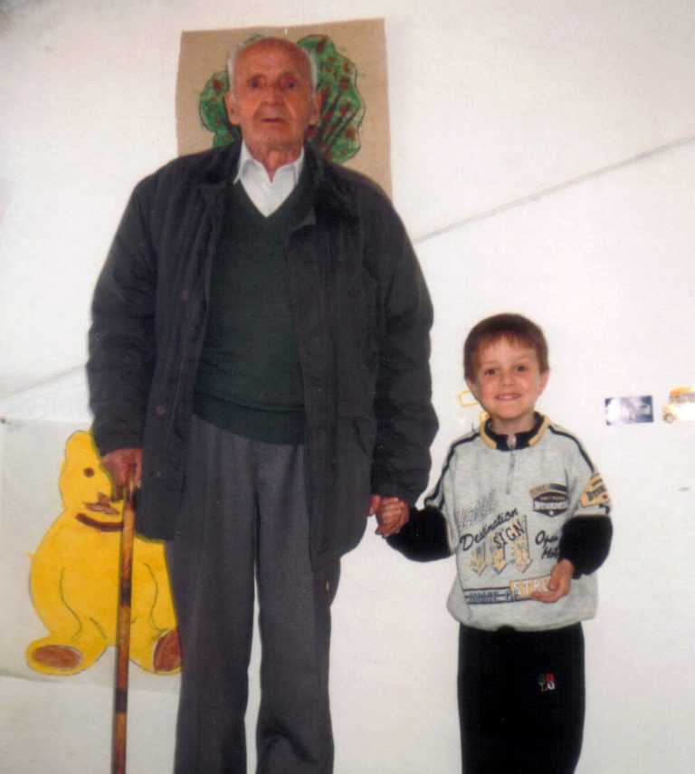 Mi abuelo y yo cogidos de la mano, posando para la foto de pie en la terraza de mi antigua casa. Detrás hay colgados algunos dibujos míos